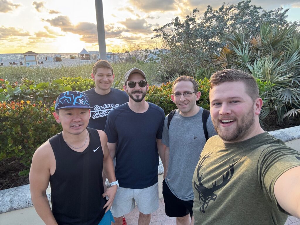 Five friends smiling outdoors near a beach at sunset.
