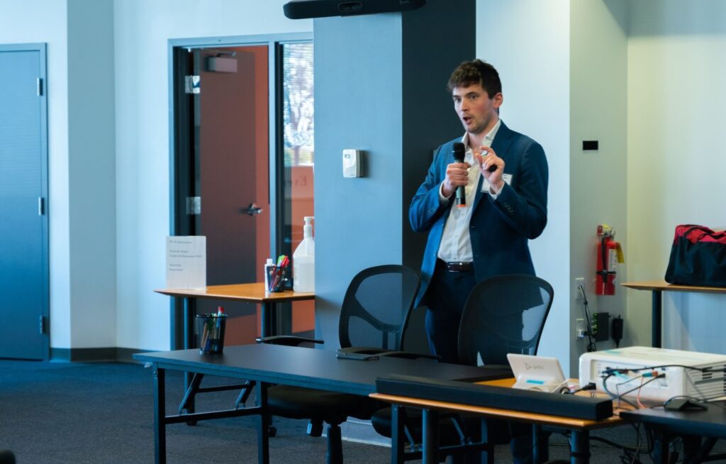 Man presenting at CFI industry event, holding a microphone and wearing a suit.